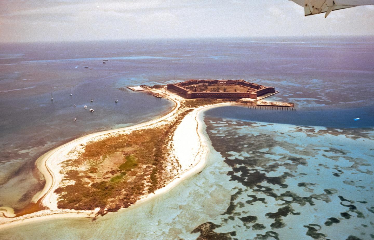 An aerial view shows a fort on a small island connected by a sandy spit to another island, surrounded by clear blue water with boats.