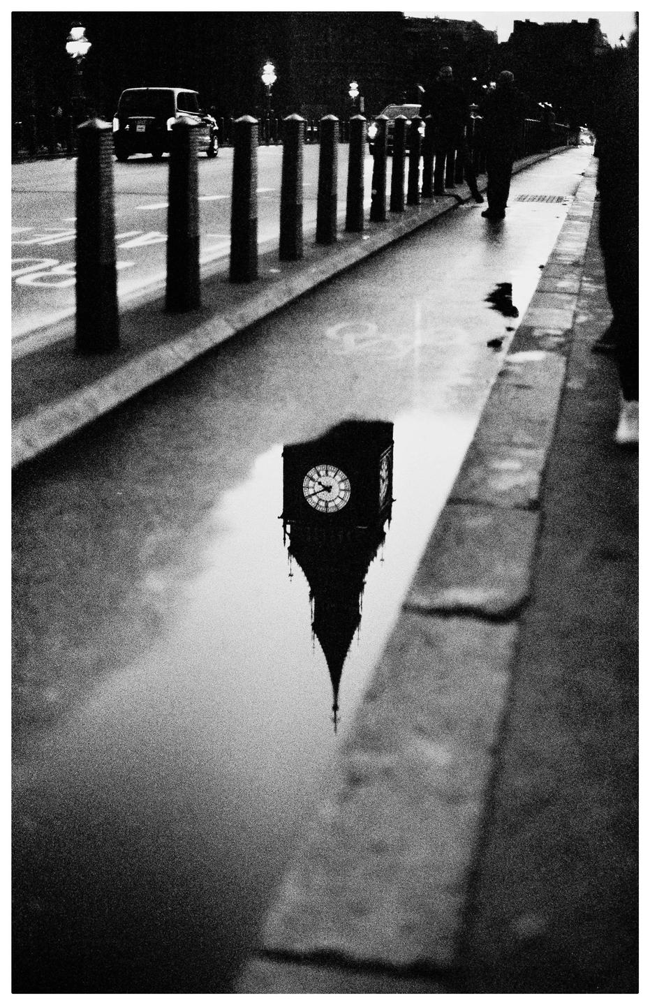 The Big Ben clock tower is reflected in a puddle on a street with bollards and a car.