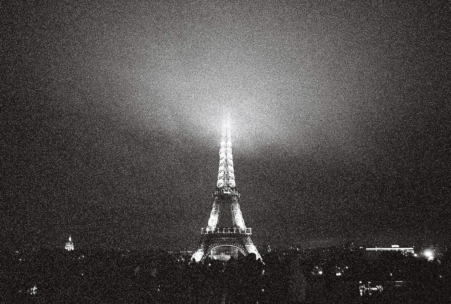 The Eiffel Tower is brightly lit at night, with a hazy sky above it and a city skyline below.