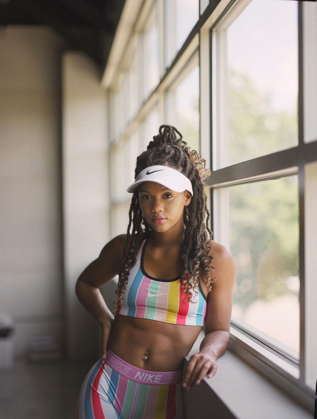 A woman with dreadlocks and a visor poses in athletic wear next to a large window.