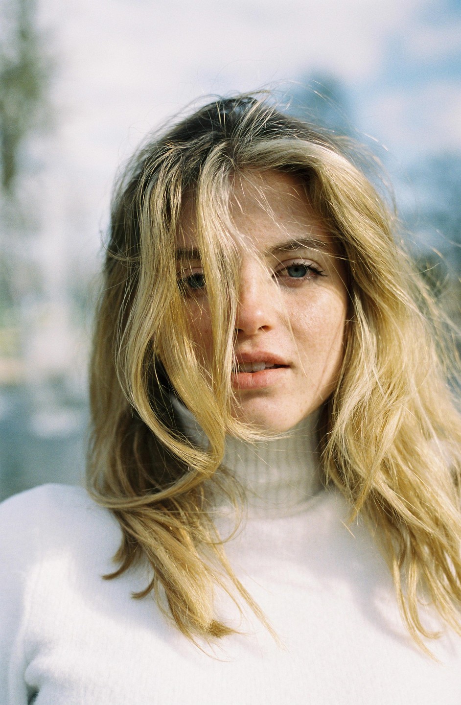 A woman with long blonde hair and a white turtleneck sweater looks directly at the viewer.