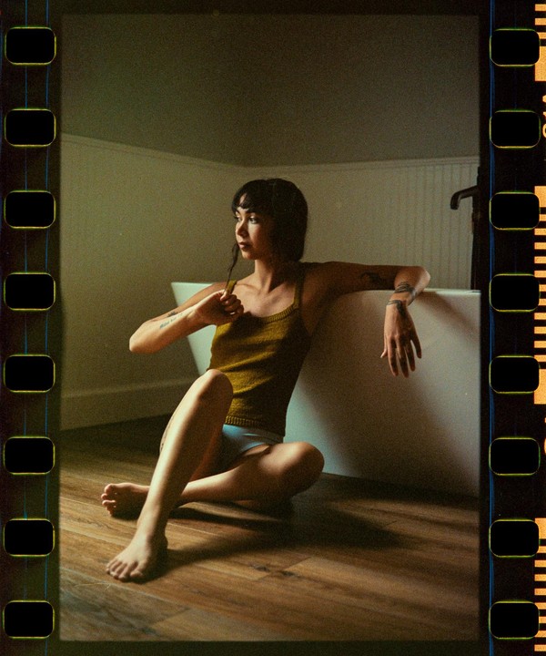 A woman sits on a wooden floor next to a bathtub, looking to her left.
