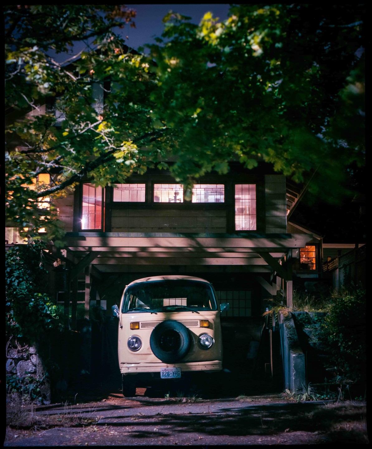 A light-colored van is parked under a house with illuminated windows at night.