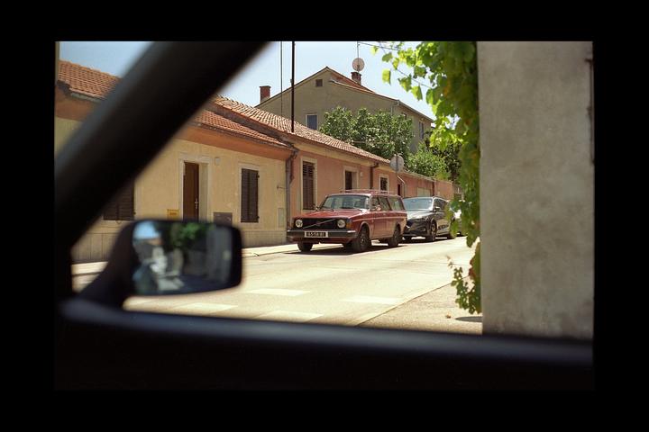 A red station wagon is parked on a street in front of buildings with terracotta roofs, viewed from inside a vehicle.
