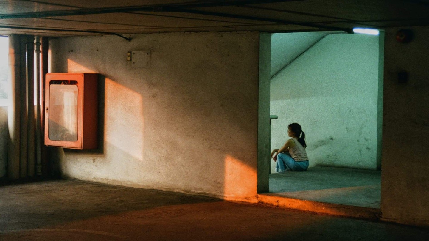 A woman sits on the floor in a dimly lit concrete room with a fire hose cabinet on the left wall.