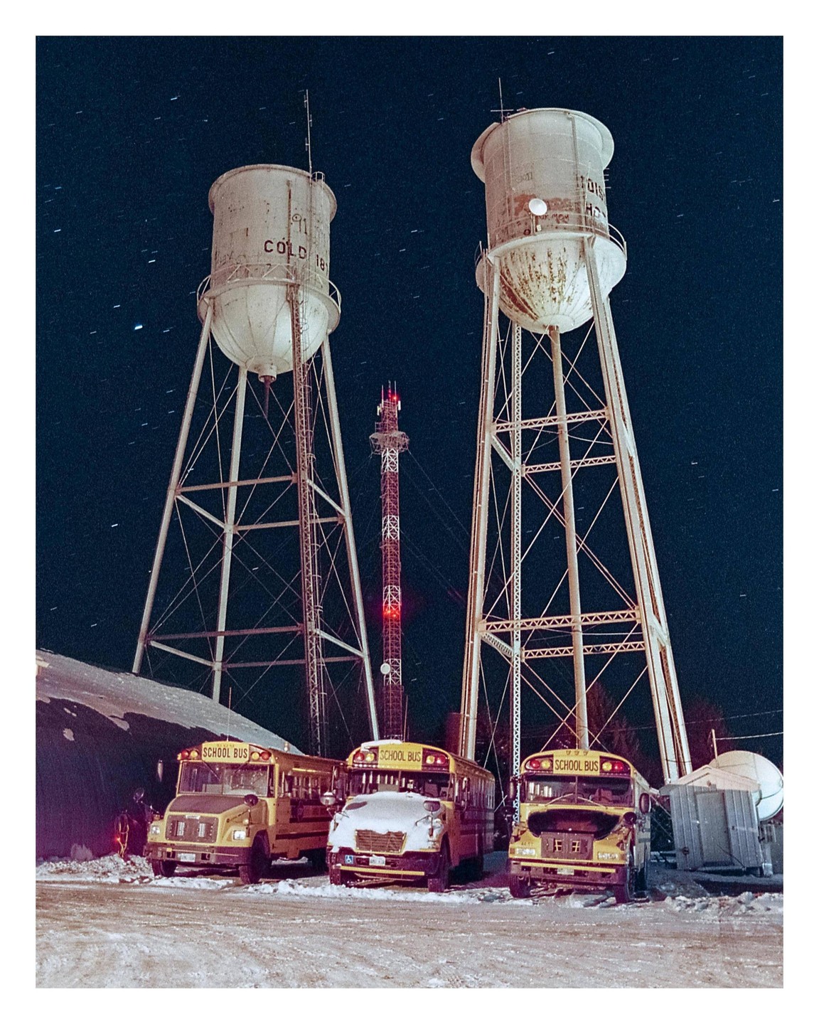 Three yellow school buses are parked in the snow beneath two water towers and a radio mast at night.