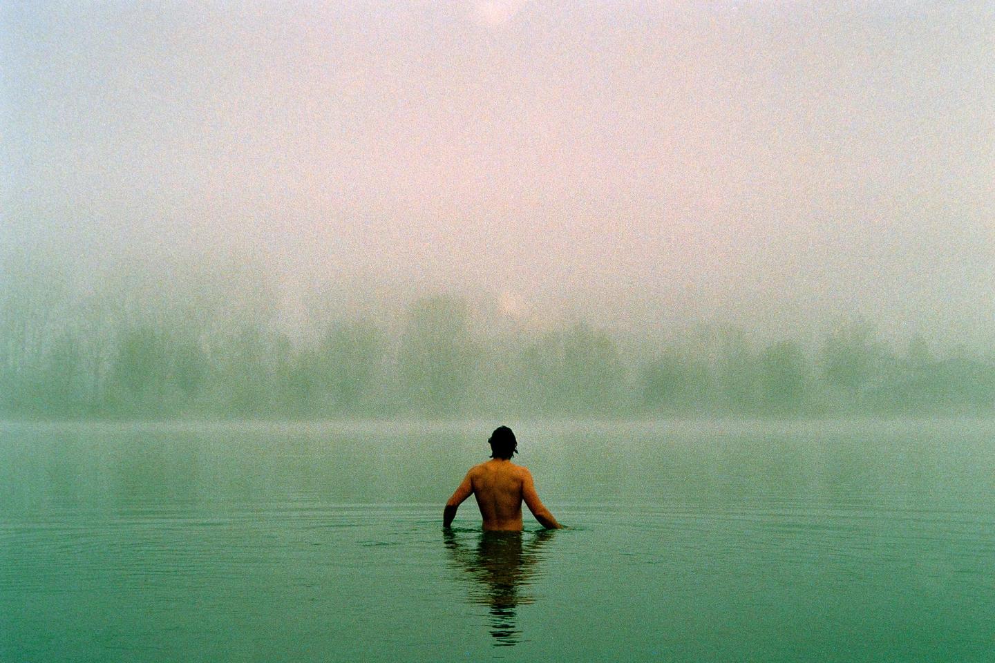 A man stands in a body of water with a hazy, tree-lined shore in the distance.