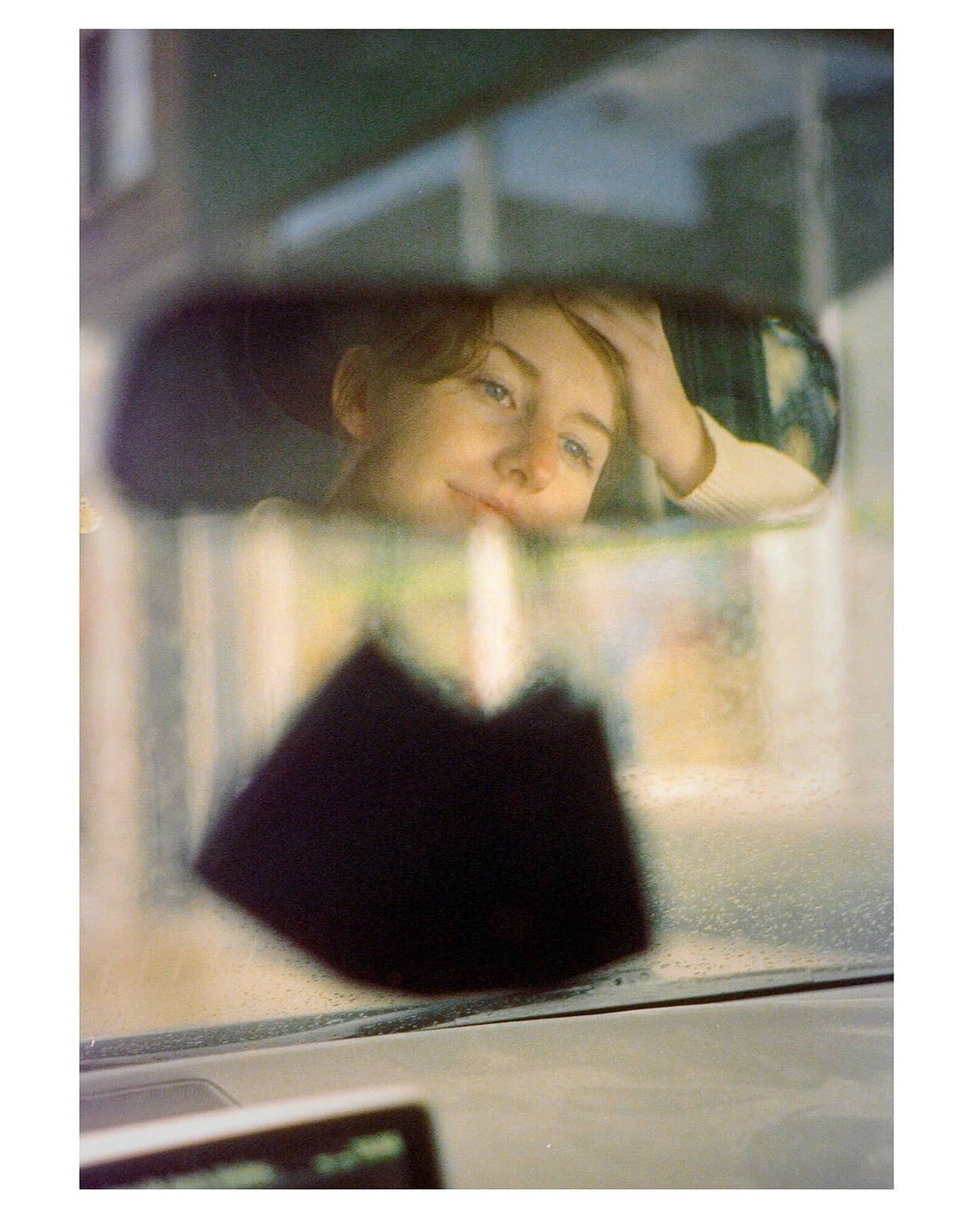 A woman is visible in a car's rearview mirror, with a fuzzy black object hanging from it.