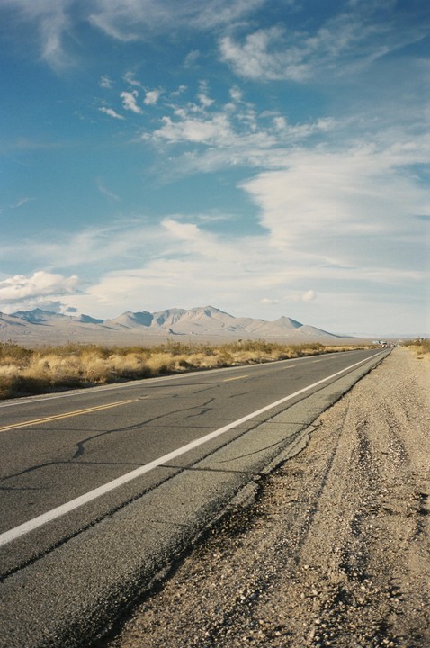 A long, empty road stretches into the distance with mountains under a blue sky with clouds.
