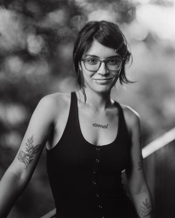 A woman with glasses and tattoos smiles at the viewer in a black and white portrait.