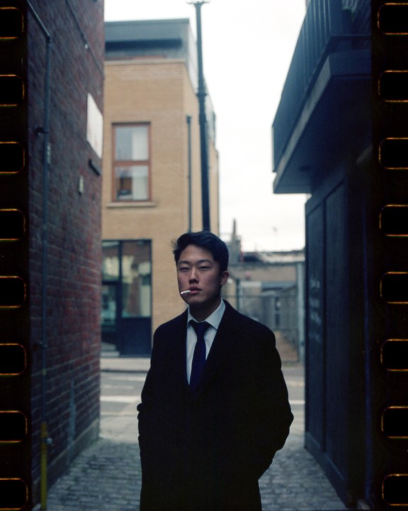 A man in a suit with a cigarette in his mouth stands in a narrow alleyway.