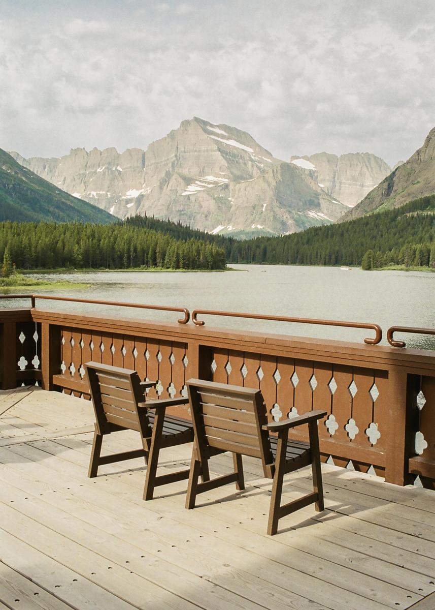 Two wooden chairs sit on a deck overlooking a lake surrounded by a forest and mountains.