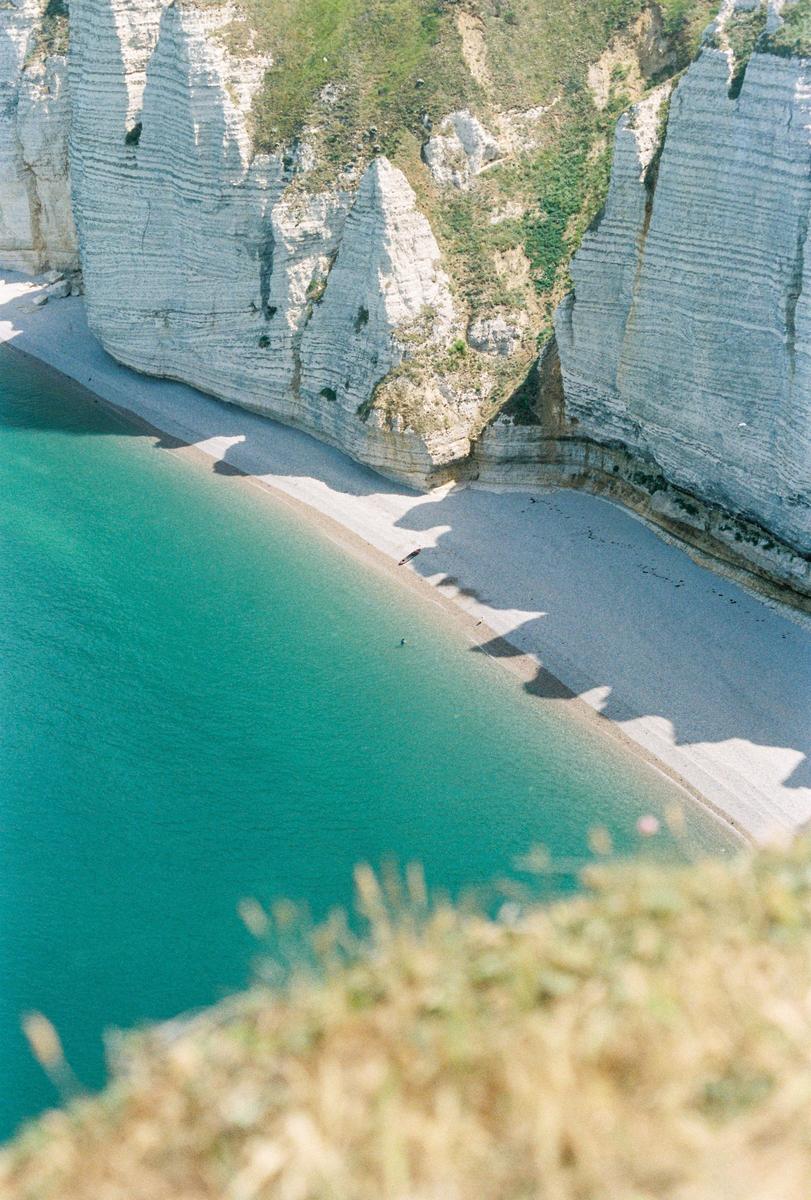 A high-angle view shows a turquoise ocean next to a pebble beach and white cliffs.