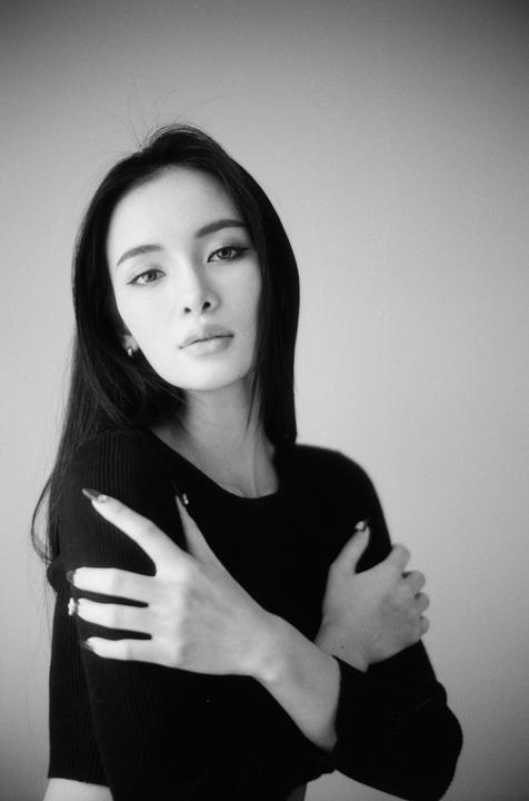 A woman with long dark hair and dark clothing looks directly at the viewer with her arms crossed.