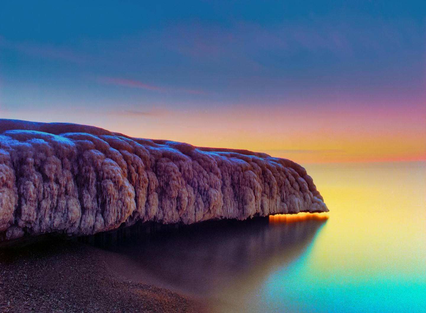 A textured rock formation overhangs calm water under a colorful sky at dusk.