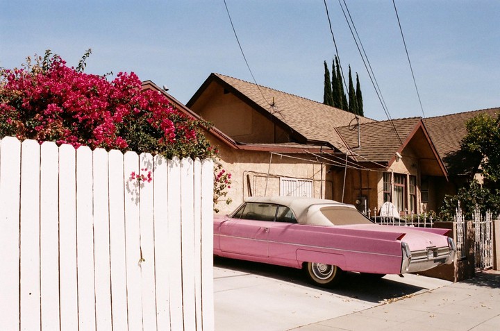 A pink convertible car is parked in a driveway next to a house with a white picket fence and pink bougainvillea.