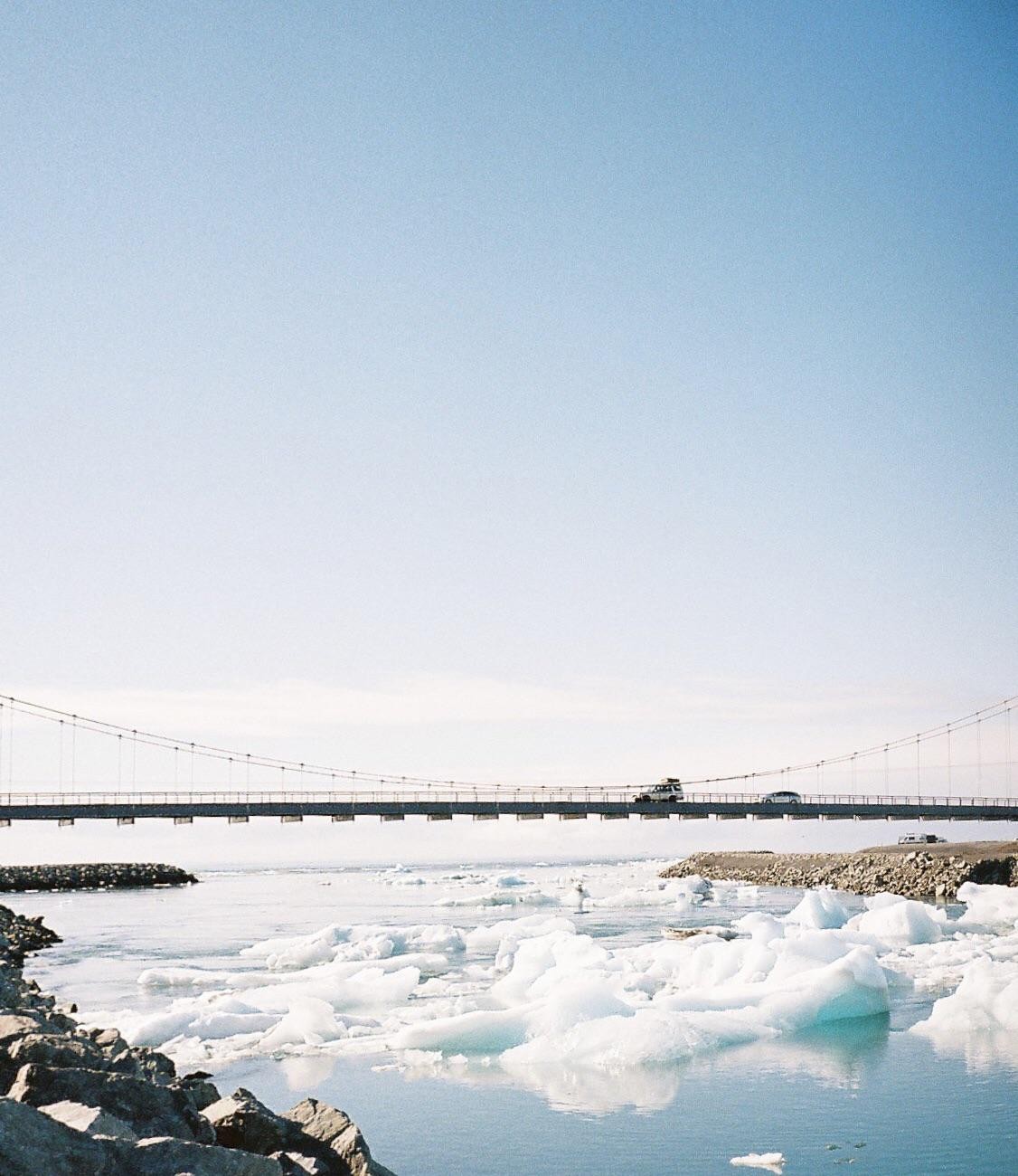 A bridge spans a body of water filled with icebergs, with a vehicle driving across the bridge.