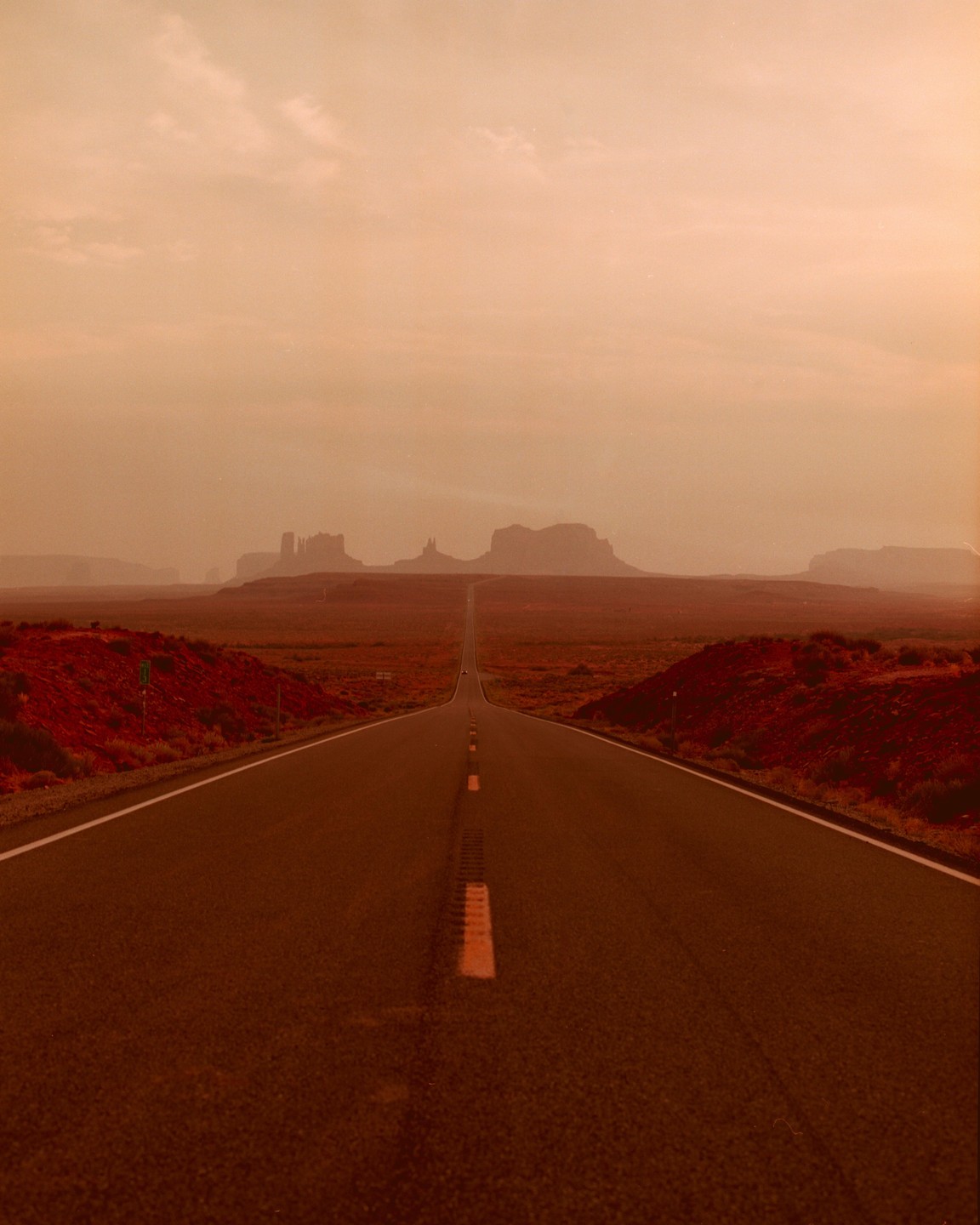 A long, straight road stretches into the distance towards a hazy horizon with distant mesas.