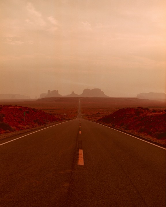 A long, straight road stretches into the distance towards a hazy horizon with distant mesas.
