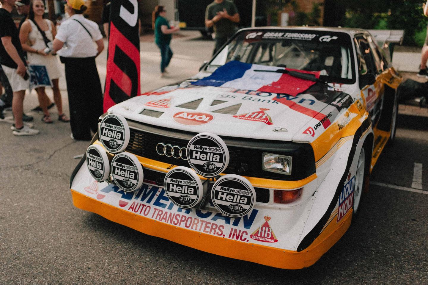 A white, orange, and black rally car with many round lights on the front is parked on asphalt with people standing around it.