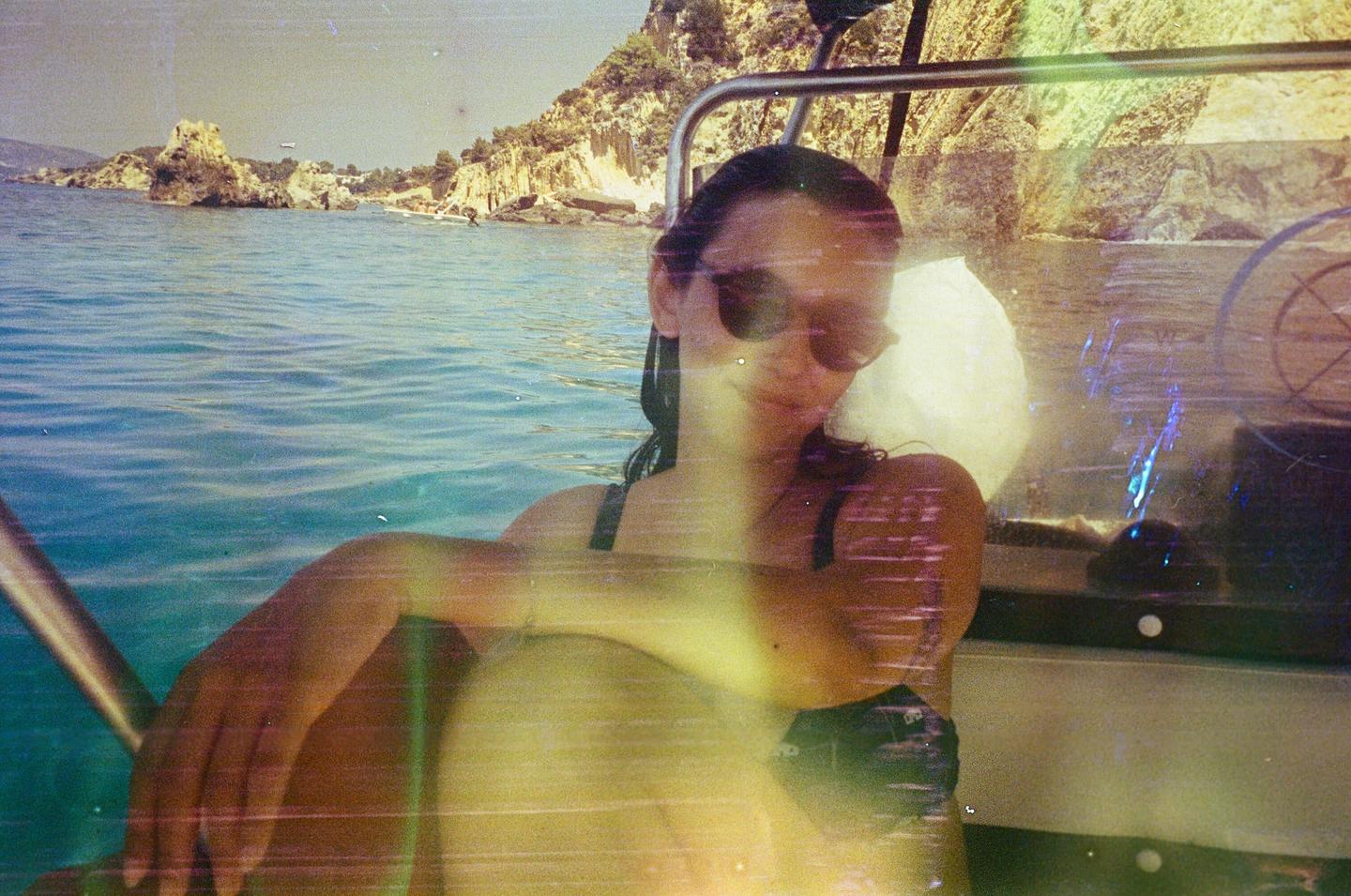 A woman wearing sunglasses sits in a boat on the water with a rocky coastline in the background.