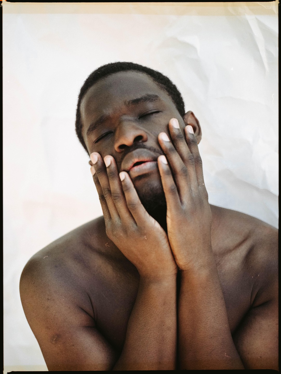 A man with closed eyes holds his hands to his face against a white background.