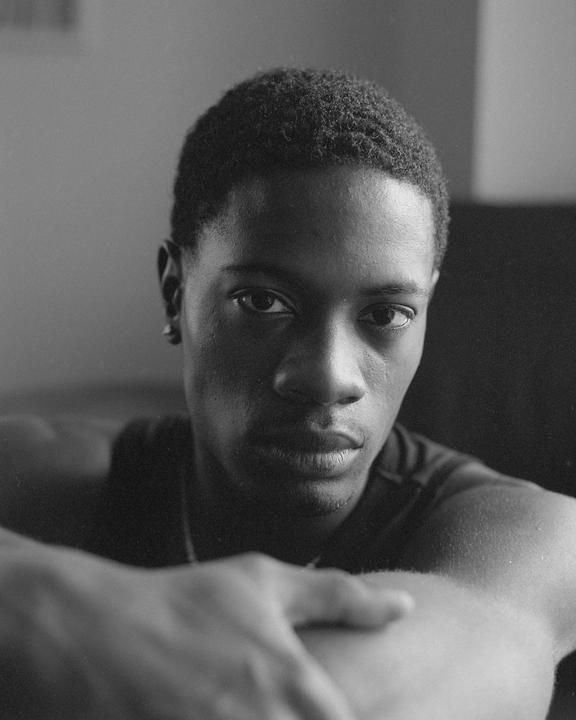 A man with short hair looks directly at the viewer, resting his arms in the foreground.
