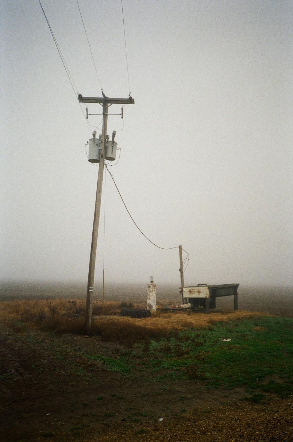 A utility pole with transformers stands in a foggy, desolate landscape next to a small structure and a dirt road.
