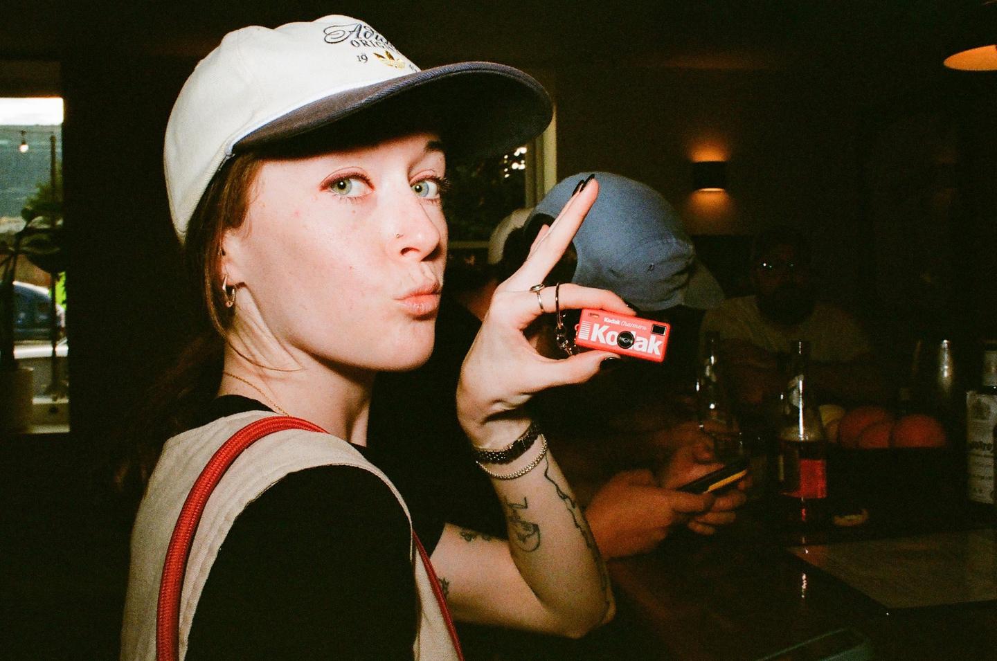 A woman wearing a cap holds up a red keychain while making a kissy face at the camera in a dimly lit bar.