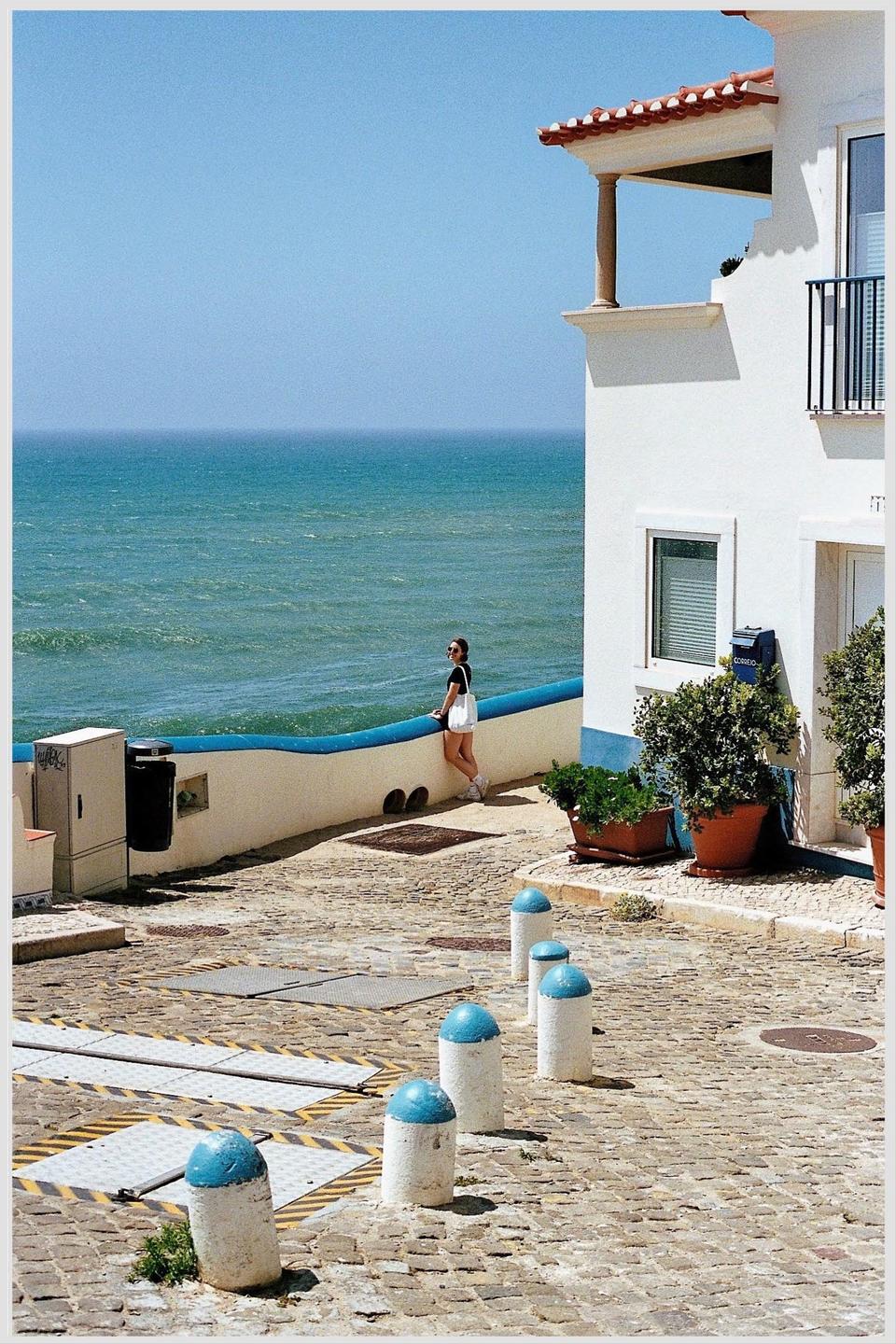 A woman stands on a cobblestone street overlooking the ocean next to a white building with a red tile roof.