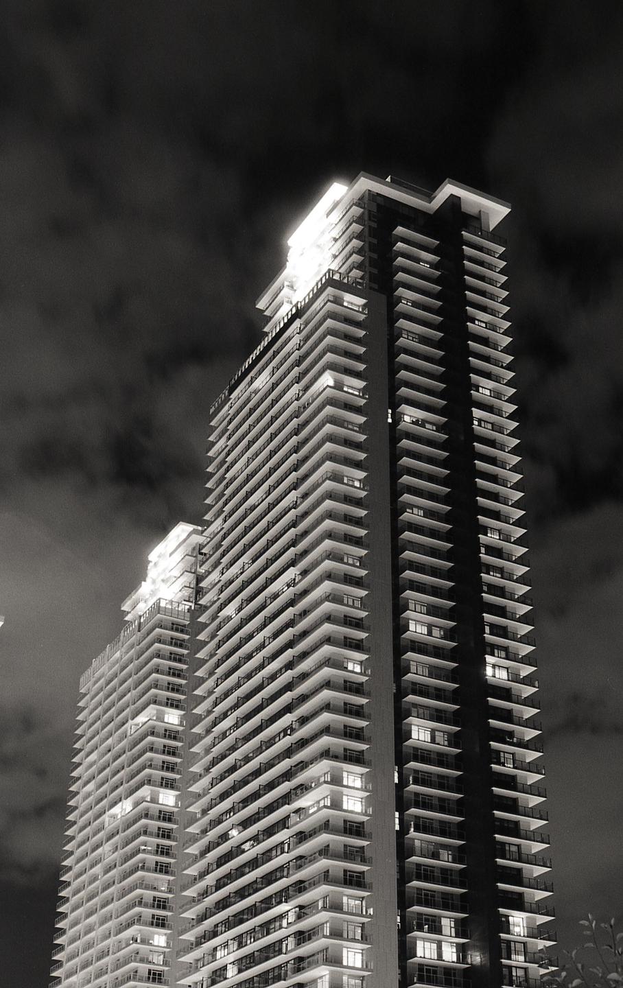 Two tall buildings with many balconies are illuminated at night against a cloudy sky.