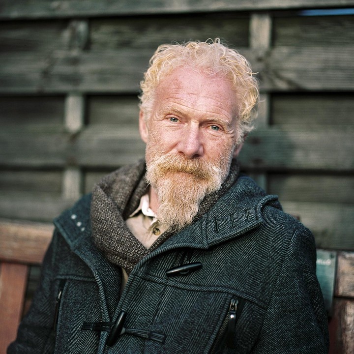 A man with a blonde beard and curly hair sits on a wooden bench in front of a wooden fence.
