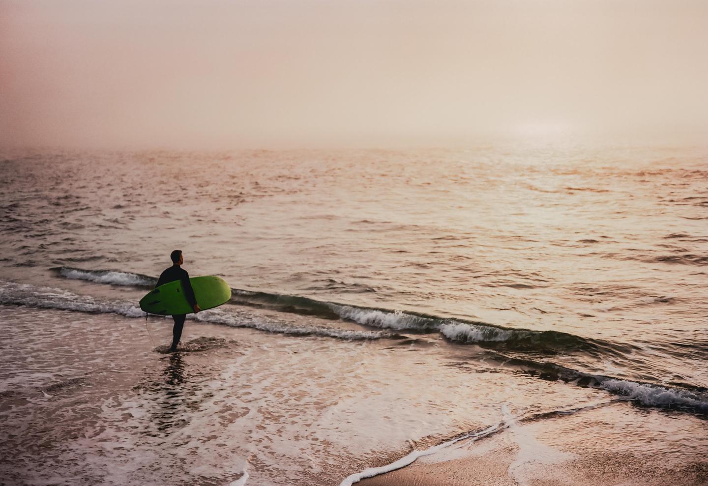 A surfer in a wetsuit stands in the shallow water of a beach, holding a green surfboard under their arm.