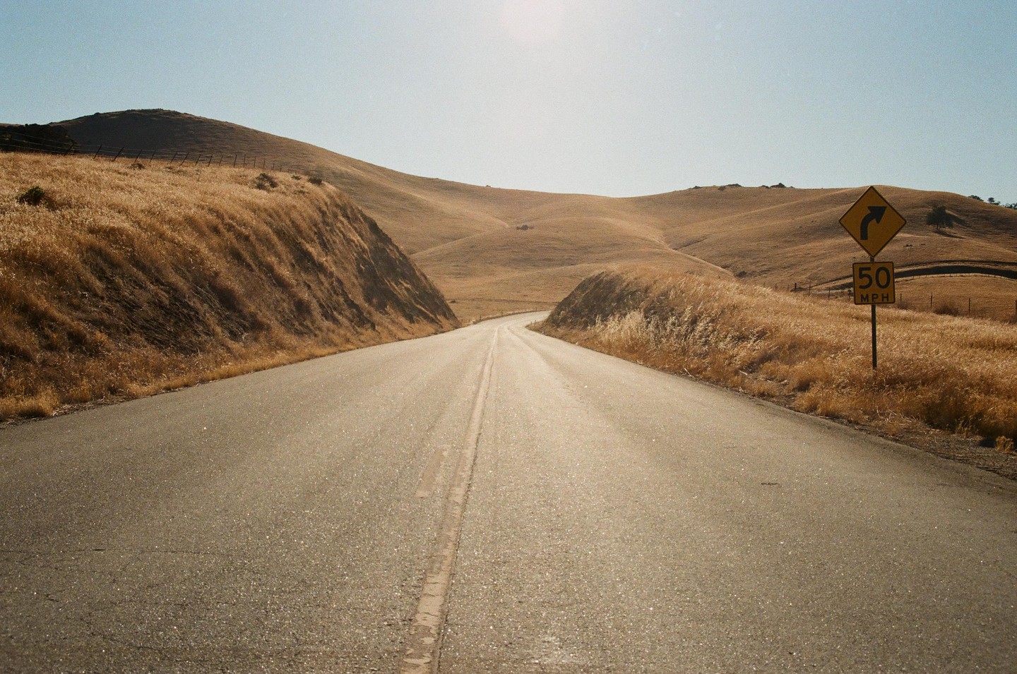 A road curves through dry, grassy hills under a bright sky.
