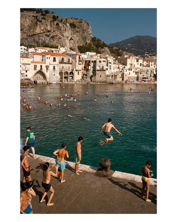 A boy jumps into the water from a concrete pier, while other boys stand on the pier and many people swim in the water in front of a town built into a cliff.