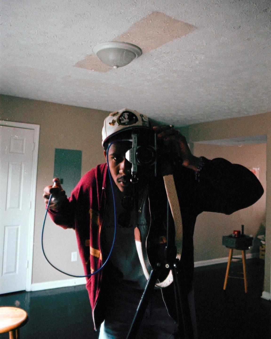 A man wearing a cap and a red jacket takes a self-portrait with a camera on a tripod.