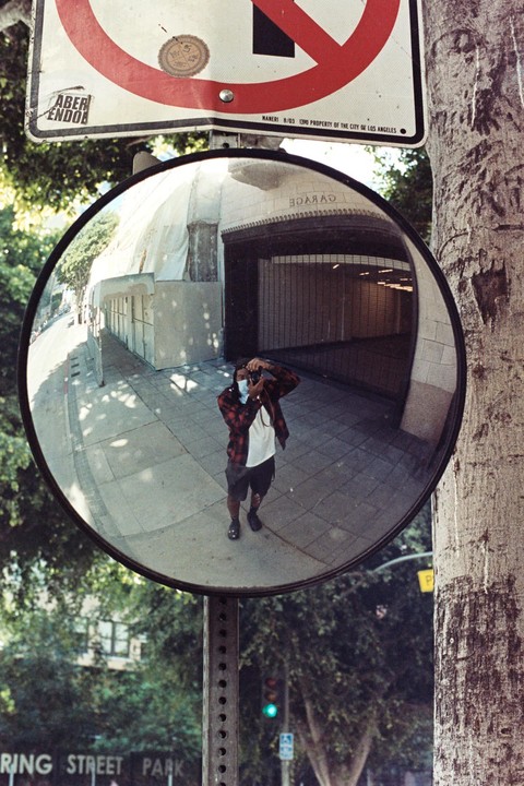A man wearing a plaid shirt and a face mask takes a self-portrait in a convex mirror attached to a pole on a street.