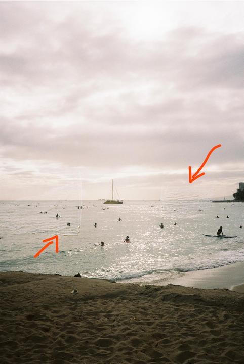 A beach scene shows many people swimming and surfing in the ocean with a boat in the distance under a cloudy sky.