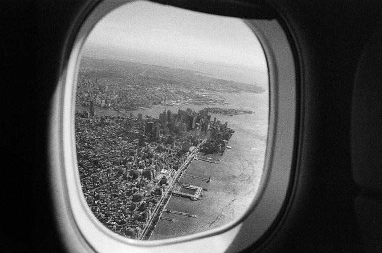 An aerial view of a city with many buildings and a body of water is seen through an airplane window.