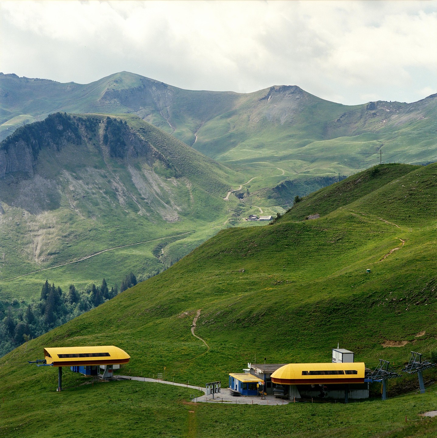 Two yellow and blue ski lift stations are visible on a green mountain slope with other mountains in the background.