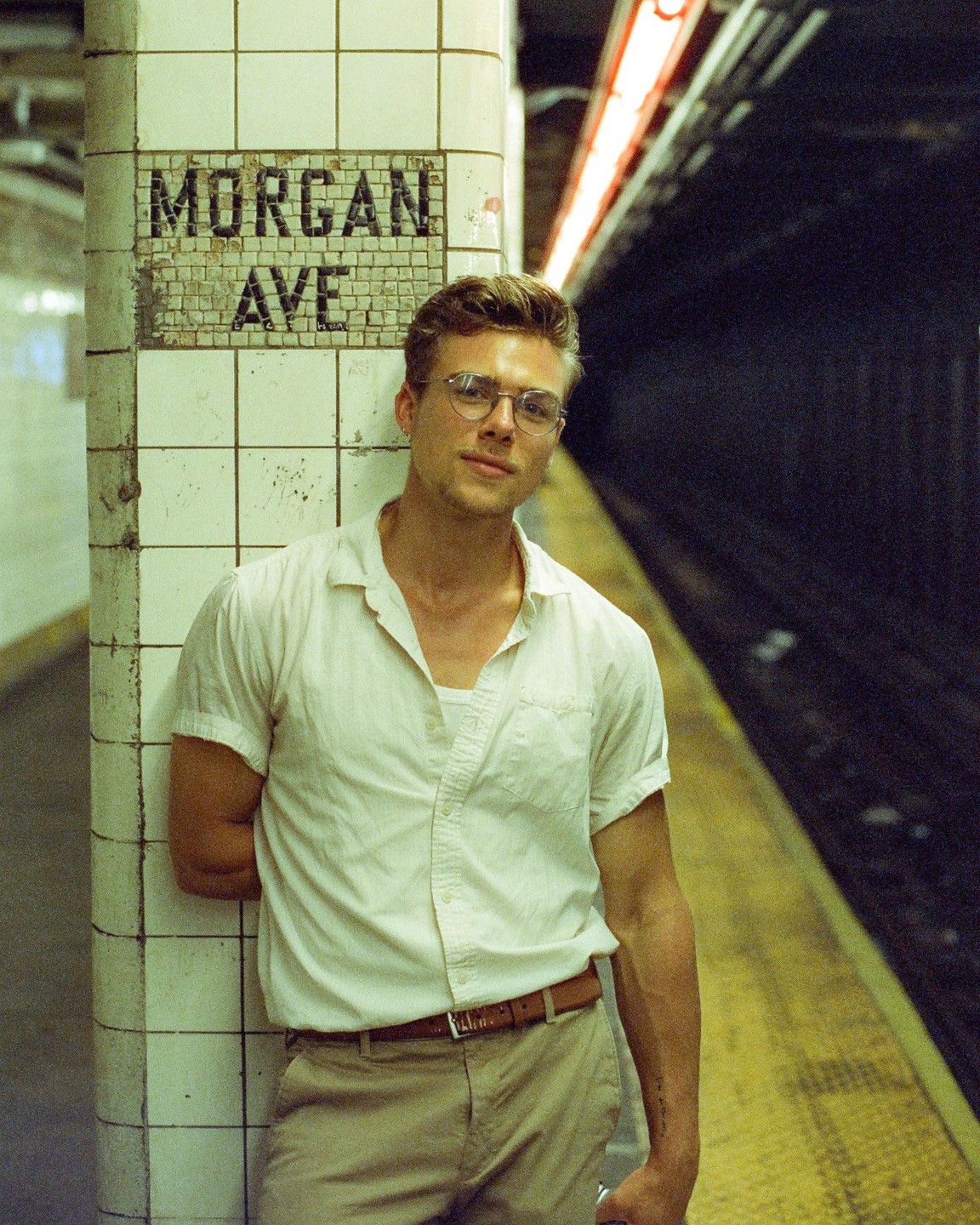 A man wearing glasses and a white shirt stands next to a tiled pillar with "MORGAN AVE" written on it in a subway station.