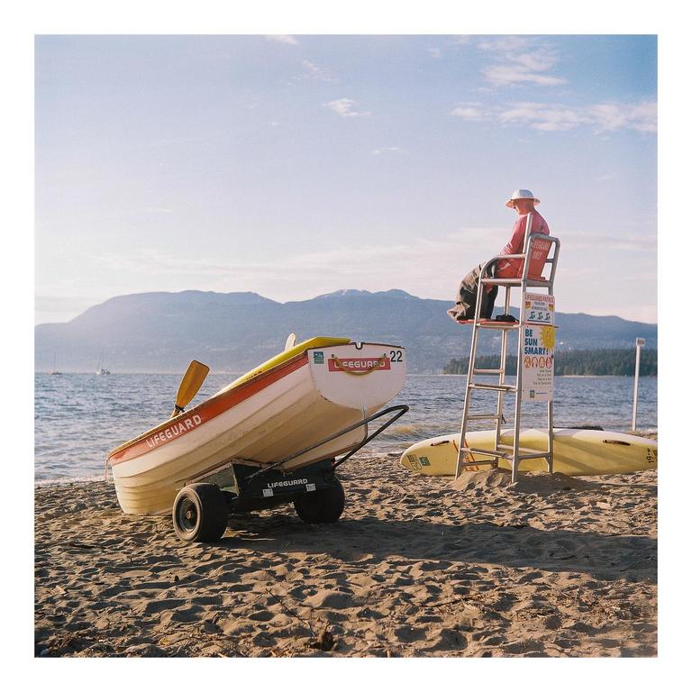 A lifeguard sits on a high chair next to a boat on a sandy beach with mountains in the background.