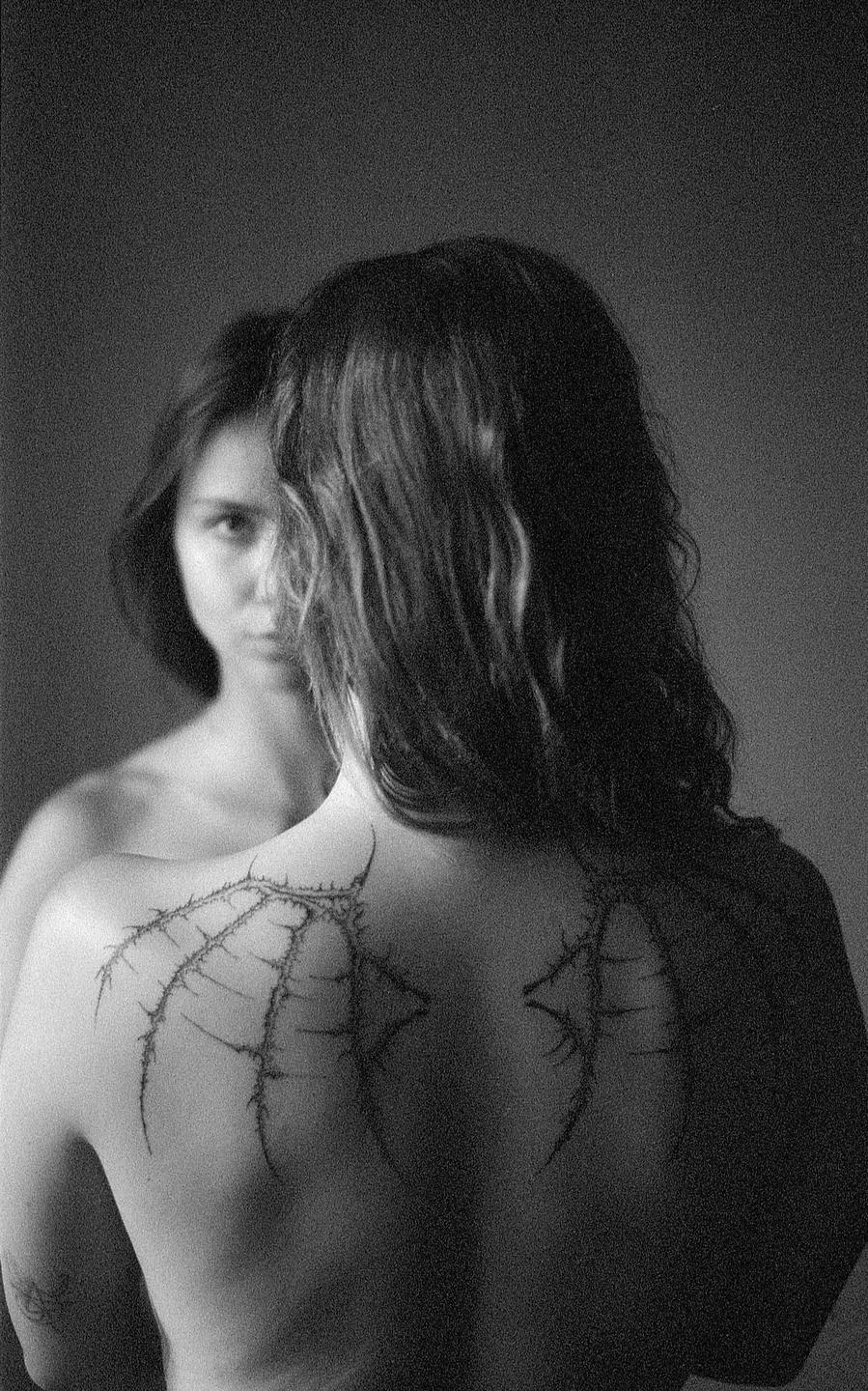 A woman with long hair and a tattoo on her back looks into a mirror, revealing her reflection looking back.