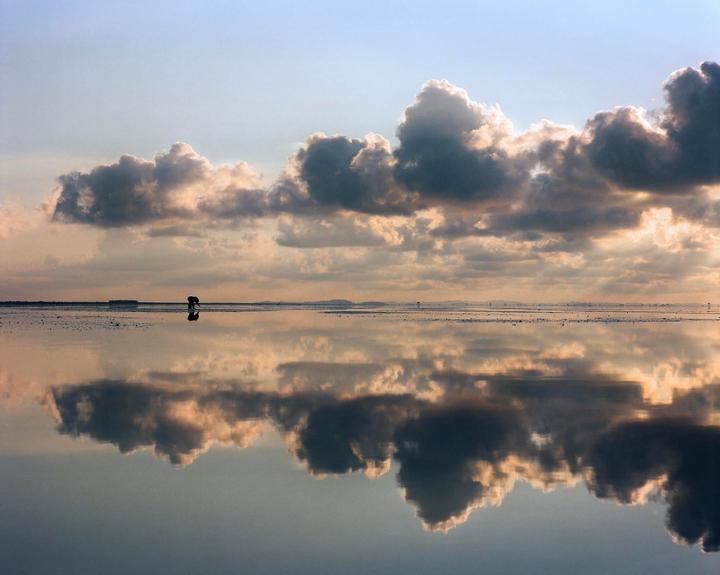 A person bends over on a reflective beach under a cloudy sky.