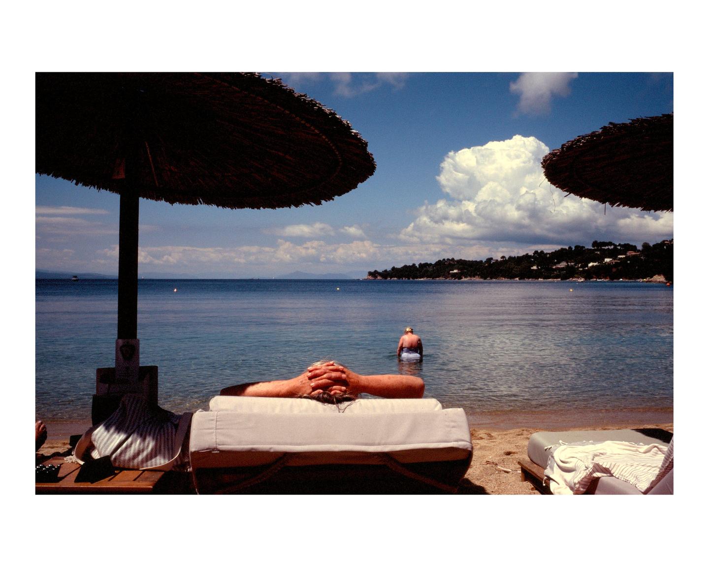 A man stands in the ocean while another person relaxes on a lounge chair under a straw umbrella on a beach.