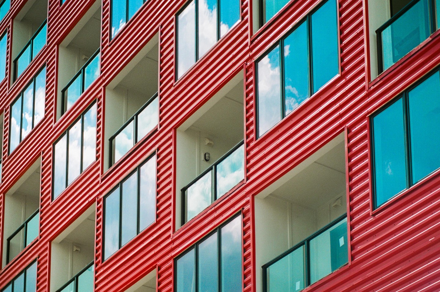 A modern building with red corrugated siding and large windows reflecting a blue sky with clouds is shown from a low angle.