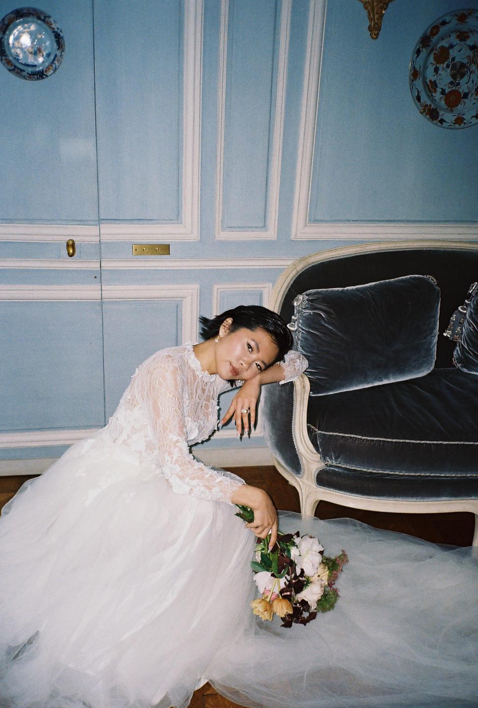 A woman in a white wedding dress is sitting on the floor next to a blue sofa, holding a bouquet of flowers.
