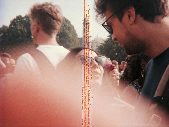 A man in sunglasses looks down at a woman in a crowd, with a vertical orange light leak down the center of the image.