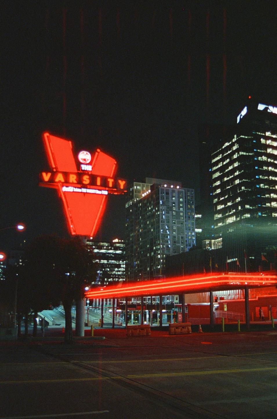 A large red neon sign for "The Varsity" stands prominently against a dark night sky, with city buildings and red light trails visible below.