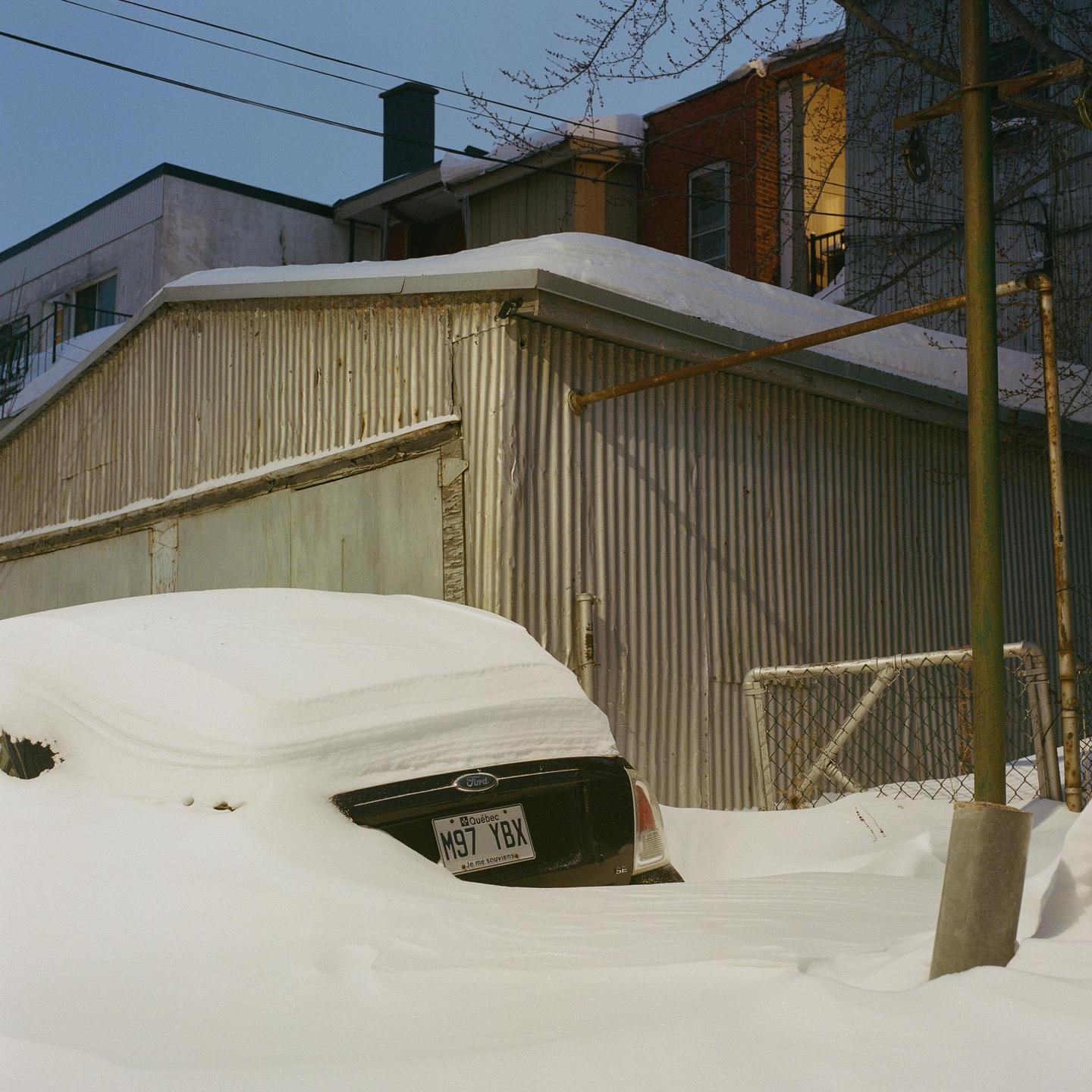 A black car is almost completely buried in deep snow next to a corrugated metal building.